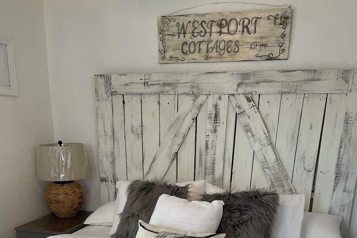 Una cama con ropa de cama blanca, un cabecero de madera rústica y un letrero que dice "WESTPORT COTTAGES EST. 1947".