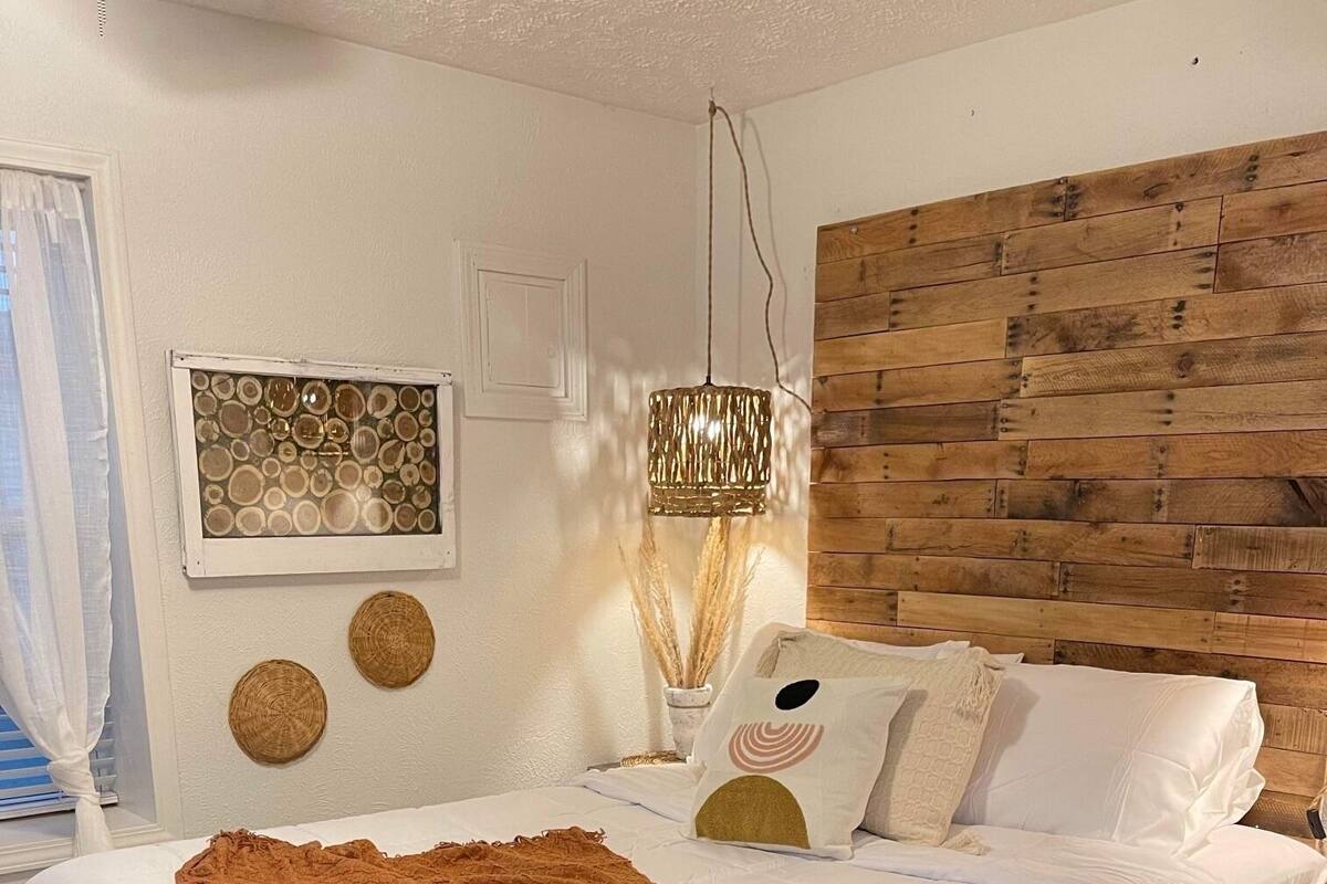 Un dormitorio con cabecera de madera, una cama grande con ropa blanca y una manta marrón, una mesita de noche y decoración en la pared, incluyendo un colgador circular y una lámpara colgante.
