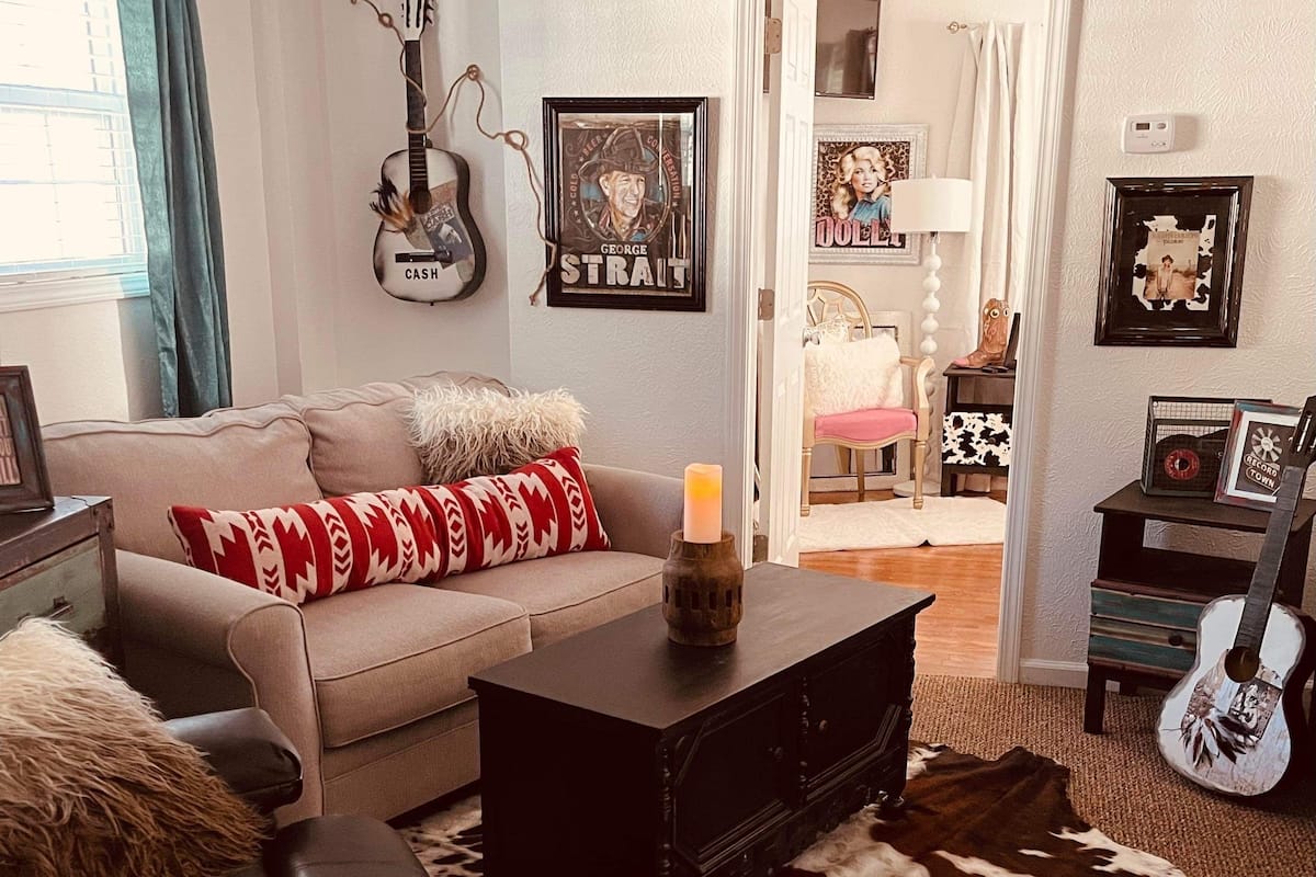Una sala de estar acogedora con un sofá, una mesa de centro y un tapete de cuero de vaca. Se observan cuadros en las paredes, una guitarra y una lámpara.