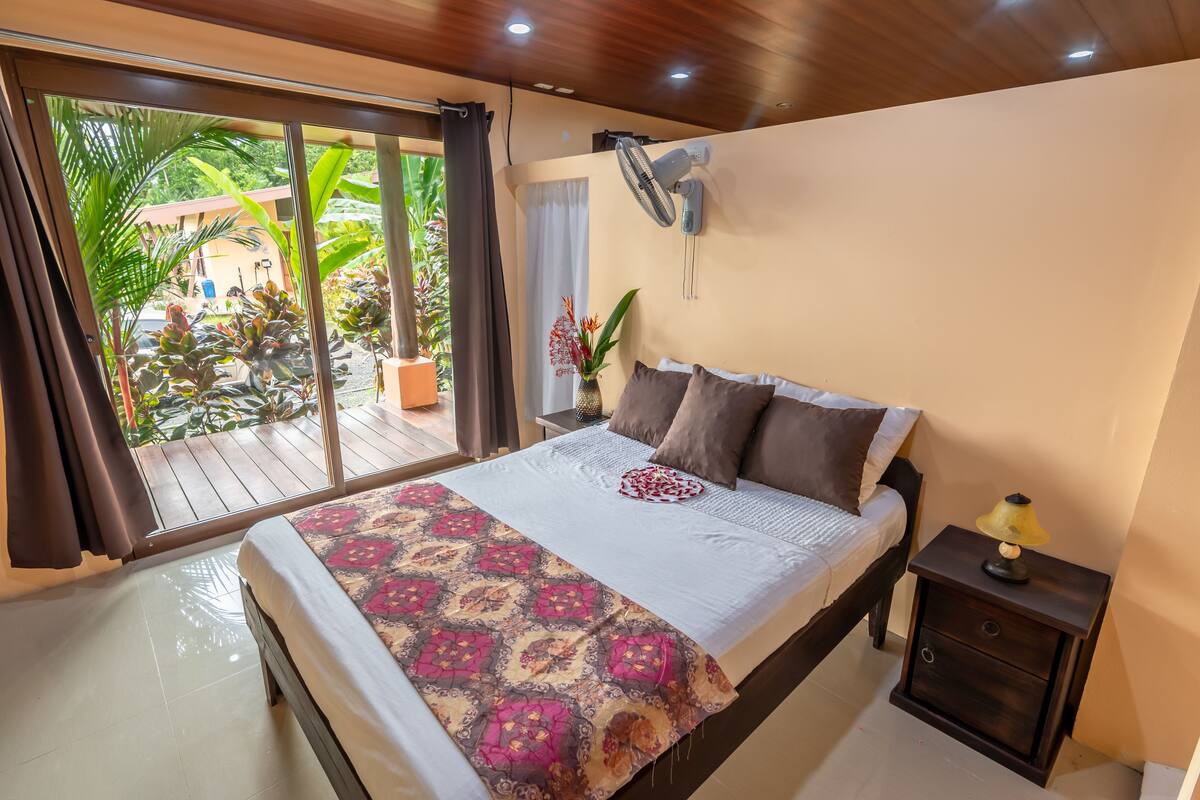 A bedroom with a bed, a nightstand, a wall-mounted fan, and a view of the outdoors.