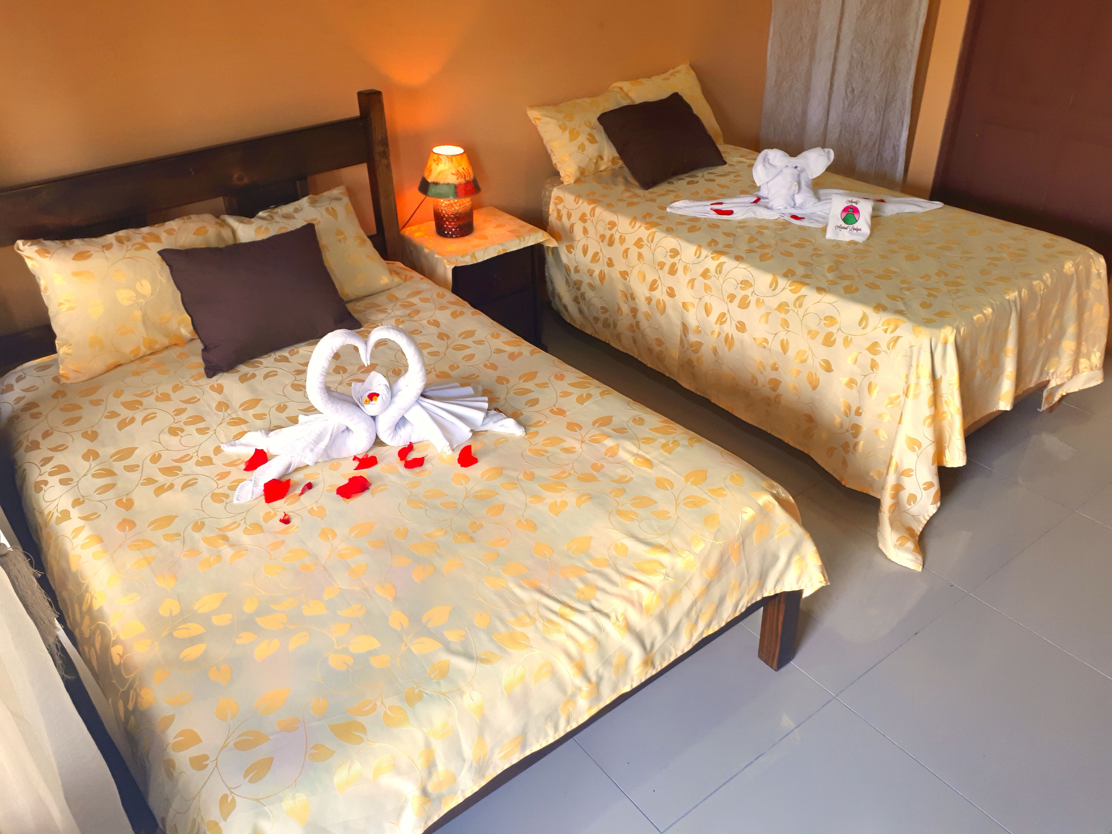 Dos camas de hotel con ropa de cama estampada con hojas, una mesita de noche con lámpara y una decoración con forma de animal hecha con toallas.
