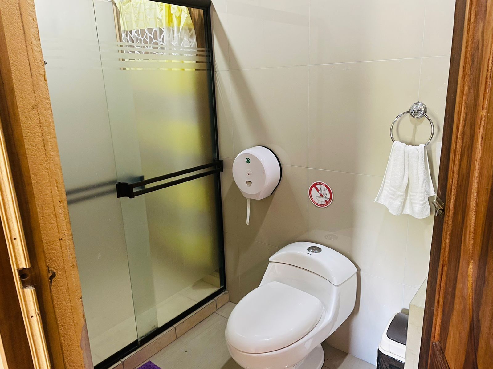 A bathroom with a glass shower enclosure, a toilet, a paper towel dispenser, and a towel rack.