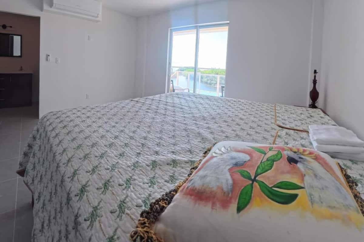 A bedroom with a large bed, a ceiling fan, a window with a view of the outdoors, and a small table with a lamp.