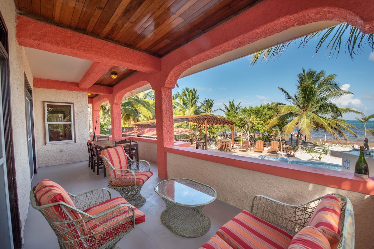 A covered outdoor seating area with wicker furniture and a glass table, overlooking a pool and palm trees.