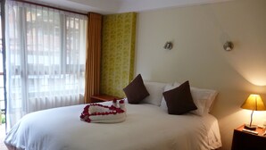A neatly made bed with a white comforter and pillows, a bedside table with a lamp, and a window with curtains. - Inka Tower (Machu Picchu)