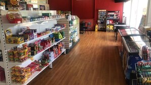 Tienda con estantes repletos de diferentes aperitivos y bebidas, mostrador de caja y vitrina con agua embotellada.