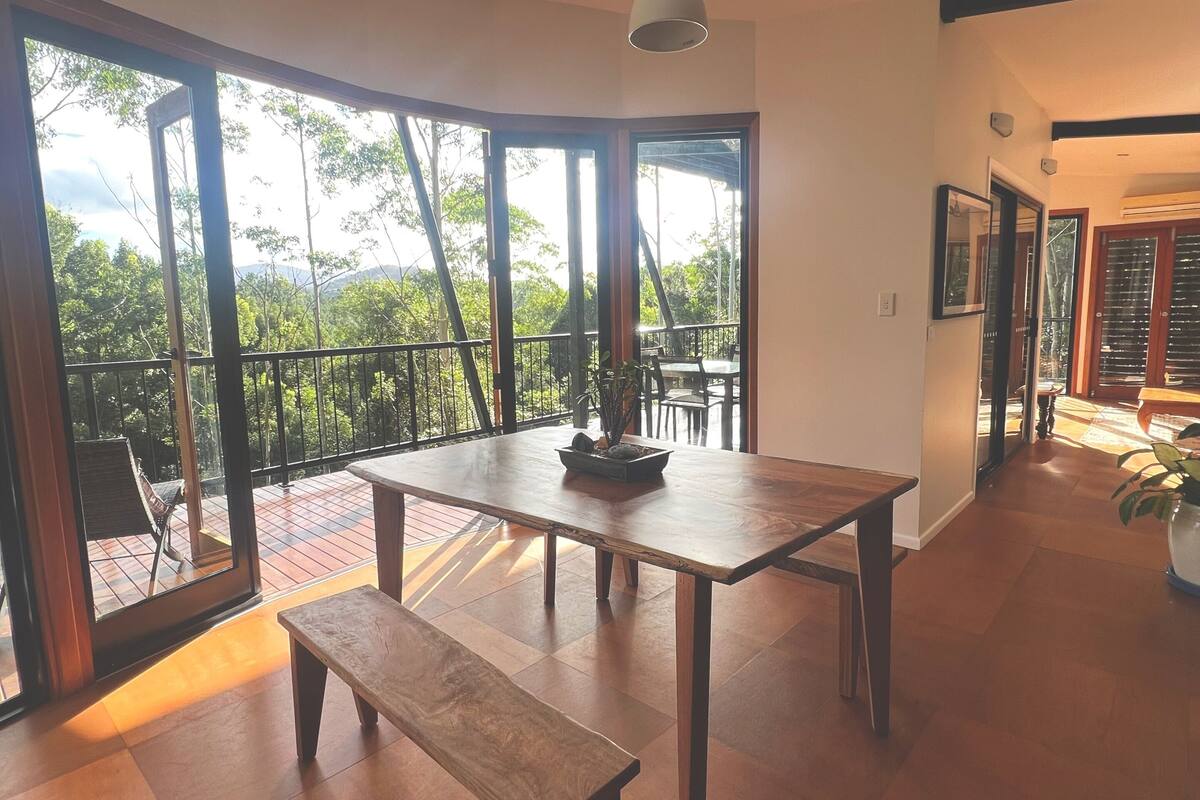 A spacious room with a large table, bench, and a view through large windows to an outdoor balcony.
