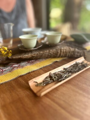 A wooden table with a woven placemat, a tray of tea leaves, and a set of green tea cups and saucers.