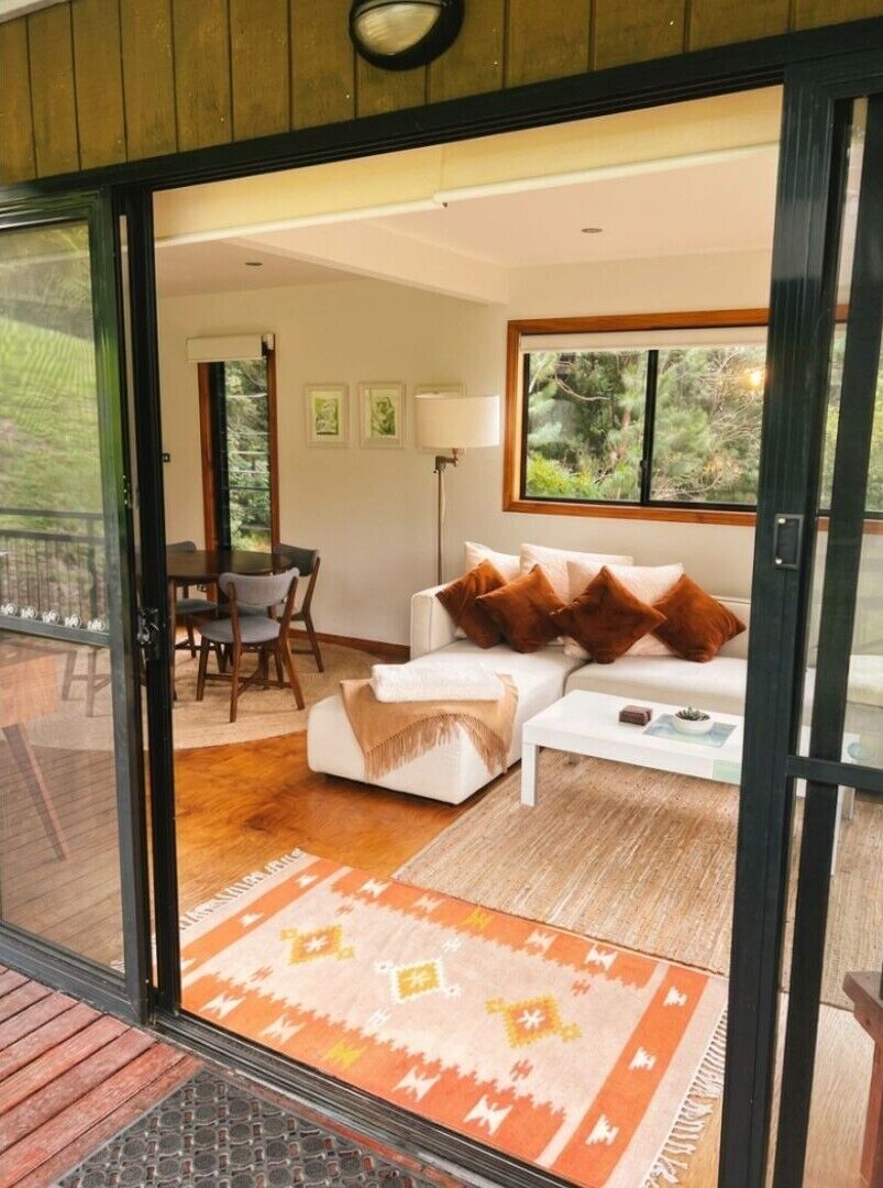 A modern living room with a sofa, coffee table, and a rug. There is a dining area with chairs and a table visible through an open door.