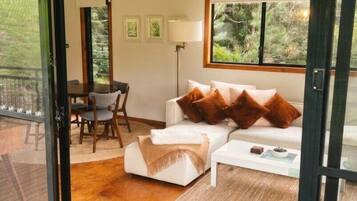 A modern living room with a sofa, coffee table, and a rug. There is a dining area with chairs and a table visible through an open door.