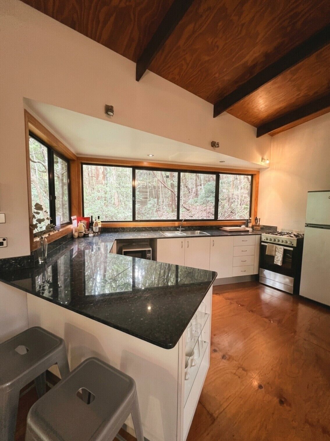 Una cocina con una isla grande, vigas de madera en el techo y vistas a un bosque a través de amplias ventanas.