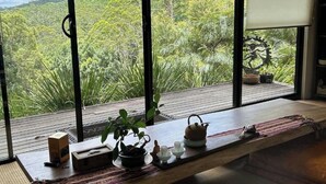 A room with a large window overlooking a forested area, a wooden table with a teapot, cups, and a box.