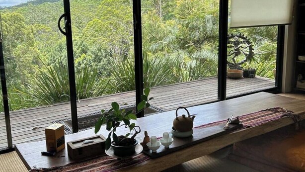 Ein Zimmer mit großem Fenster mit Blick auf eine bewaldete Landschaft, ein Holztisch mit Teekanne, Tassen und einer Schachtel.