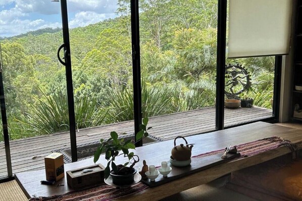 A room with a large window overlooking a forested area, a wooden table with a teapot, cups, and a box.