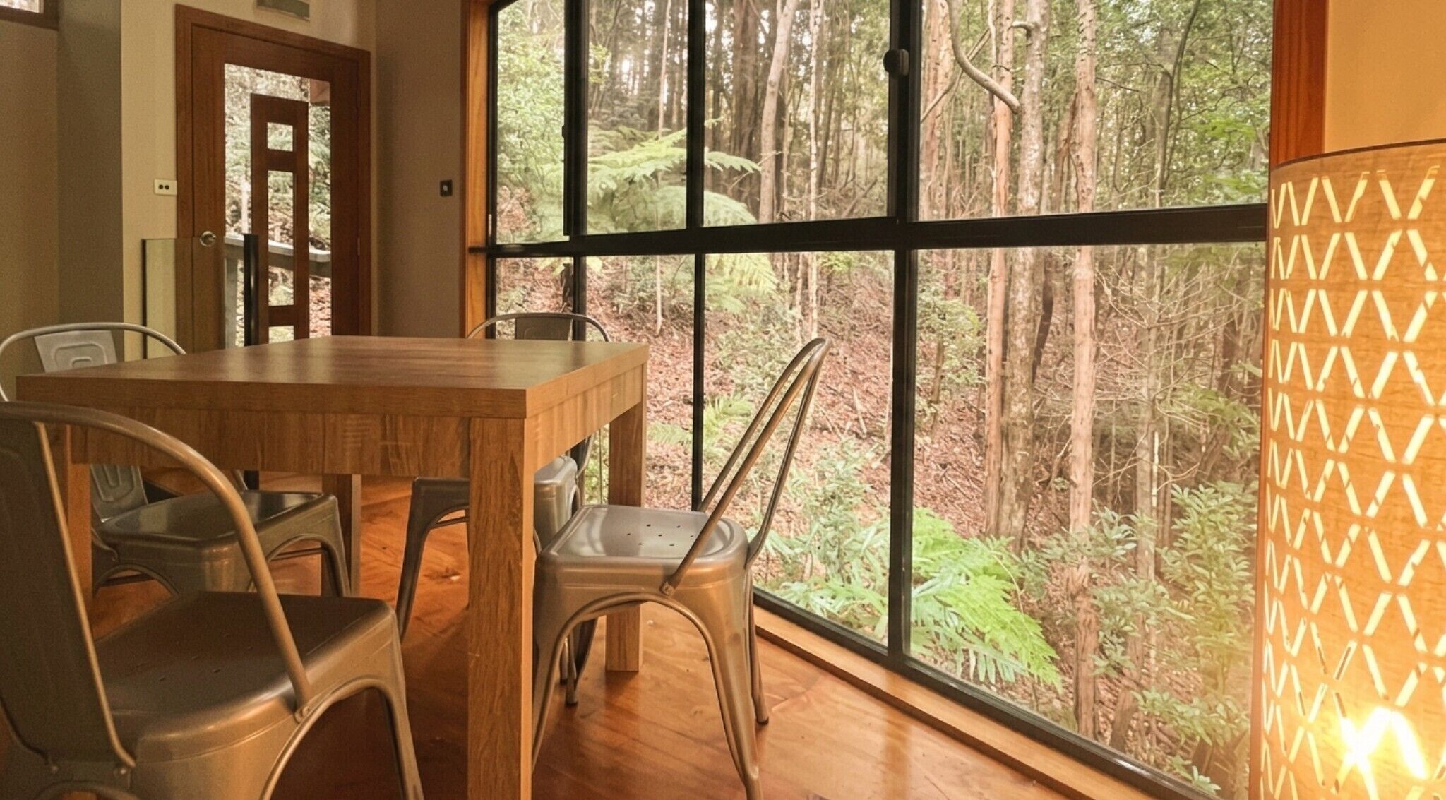 A dining area with a wooden table and metal chairs, large windows with a forest view, and a wall-mounted lamp.