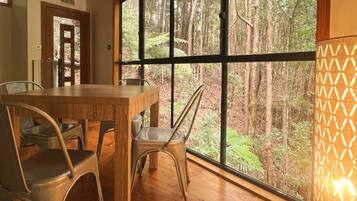 A dining area with a wooden table and metal chairs, large windows with a forest view, and a wall-mounted lamp.