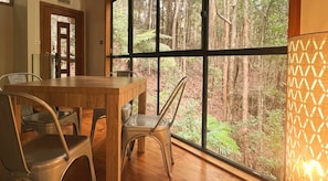 A dining area with a wooden table and metal chairs, large windows with a forest view, and a wall-mounted lamp. - Ecoasis Mami Wata Retreat (Smiths Creek)