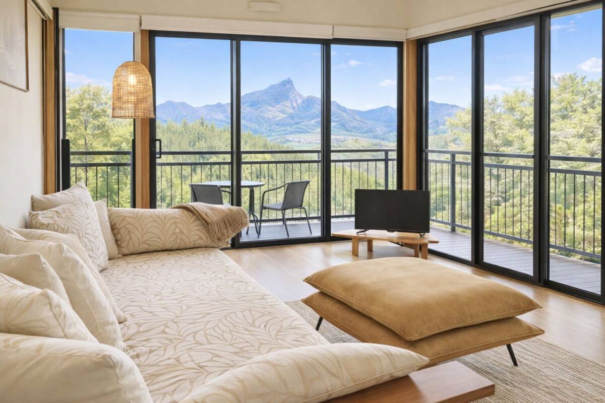 A modern living room with a large sofa, a flat-screen TV, and a balcony with a mountain view.