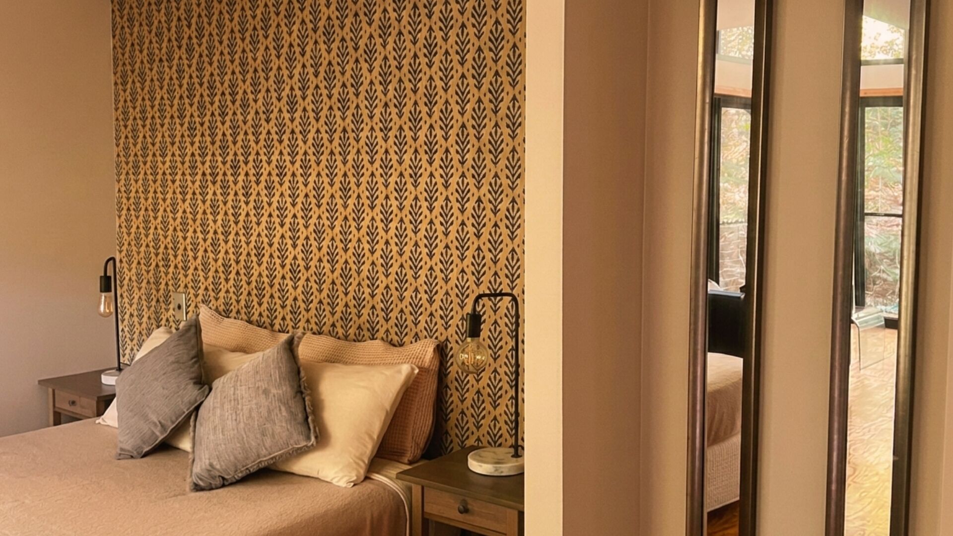 A bedroom with a patterned wallpaper, a bed with pillows, a bedside table with a lamp, and a view through a window.