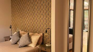 A bedroom with a patterned wallpaper, a bed with pillows, a bedside table with a lamp, and a view through a window.