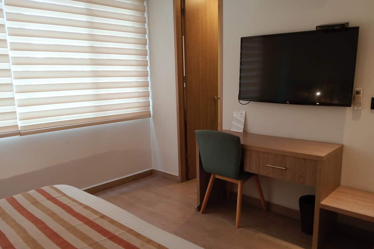 Un dormitorio con cama, escritorio, silla, televisor y ventana con persianas.