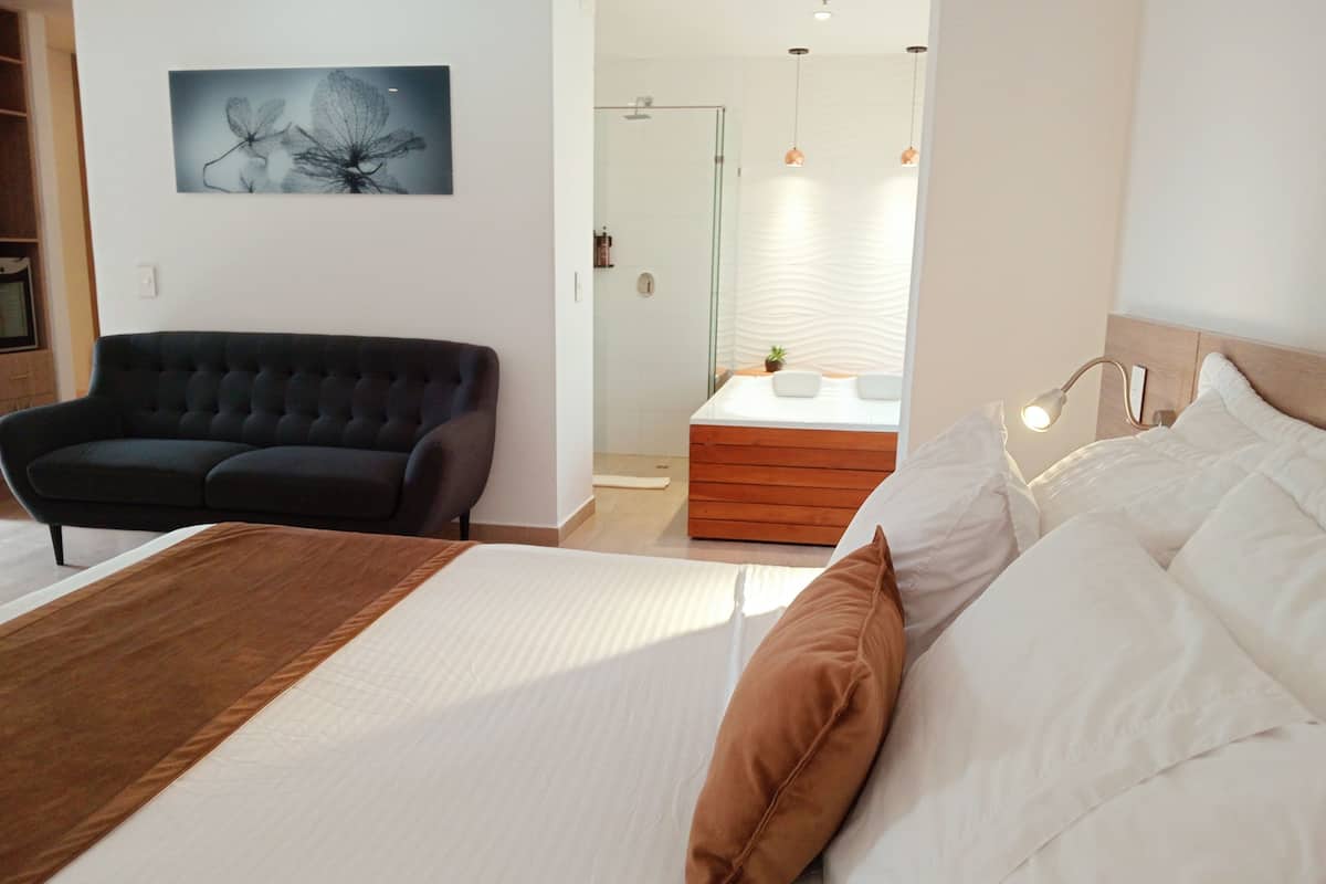 Un dormitorio moderno con una cama grande, un sofá negro y un baño pequeño visible a través de una puerta abierta.
