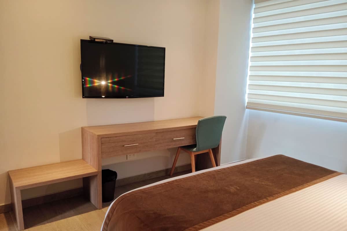 Un dormitorio con una cama, un escritorio de madera, una silla y un televisor colocado en la pared.