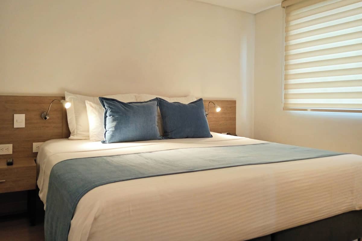 Una cama bien hecha con cabecera de madera, dos lámparas de noche y una ventana con persianas.