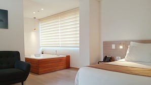 A modern hotel room with a large bed, a separate bathroom with a bathtub, a sofa, and a large window with blinds. - Hotel Lomas 10 (Medellín)