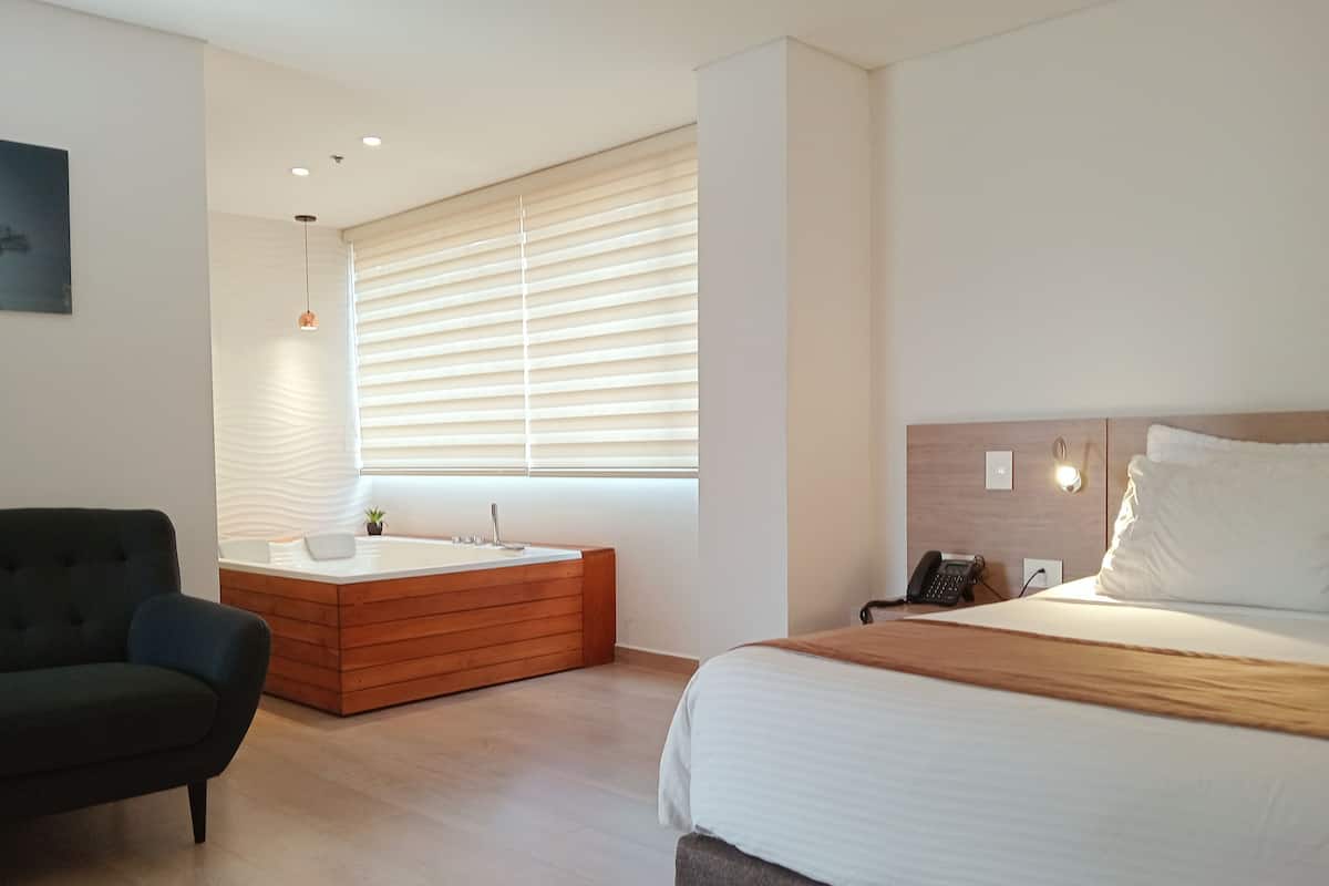 Una habitación de hotel moderna con una cama grande, un baño independiente con bañera, un sofá y un amplio ventanal con persianas.
