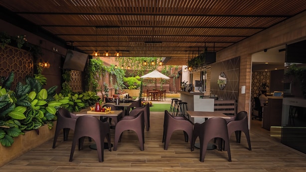 An outdoor dining area with wooden flooring, purple chairs, a central table with a white umbrella, and a bar area with a sign reading "BAR". - Hotel Lomas 10 (Medellín)