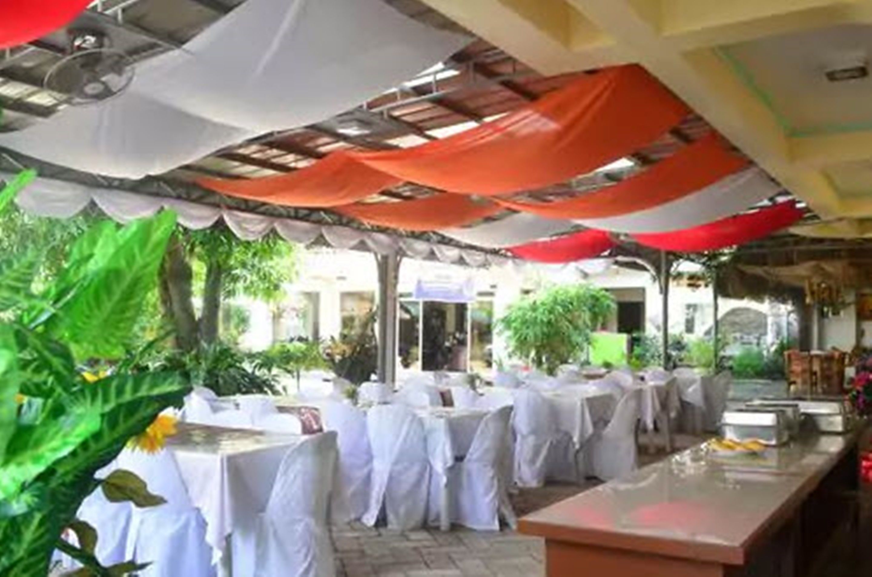 An outdoor dining area with tables covered in white tablecloths and chairs, surrounded by greenery and a covered structure with orange and white drapes.
