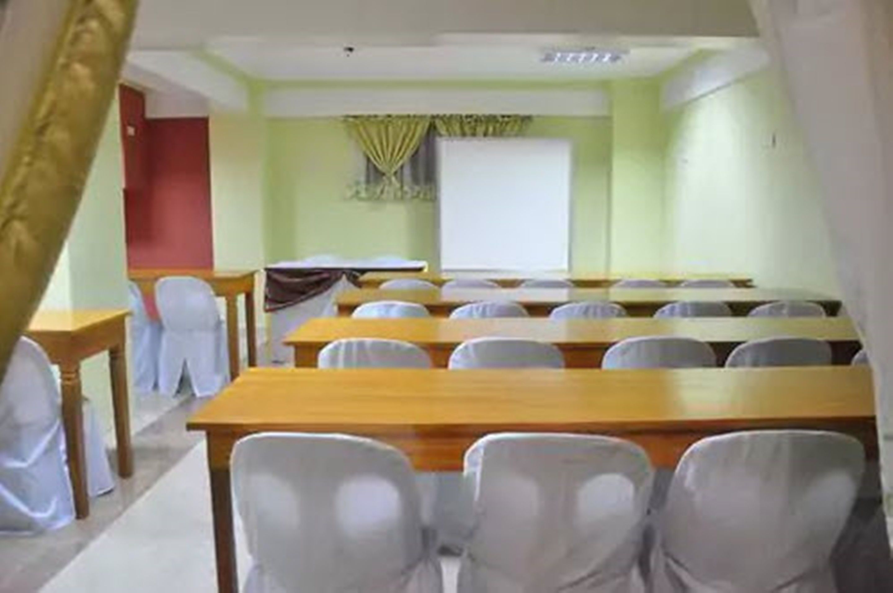 A conference room with rows of tables and chairs, a projector screen, and a window with curtains.