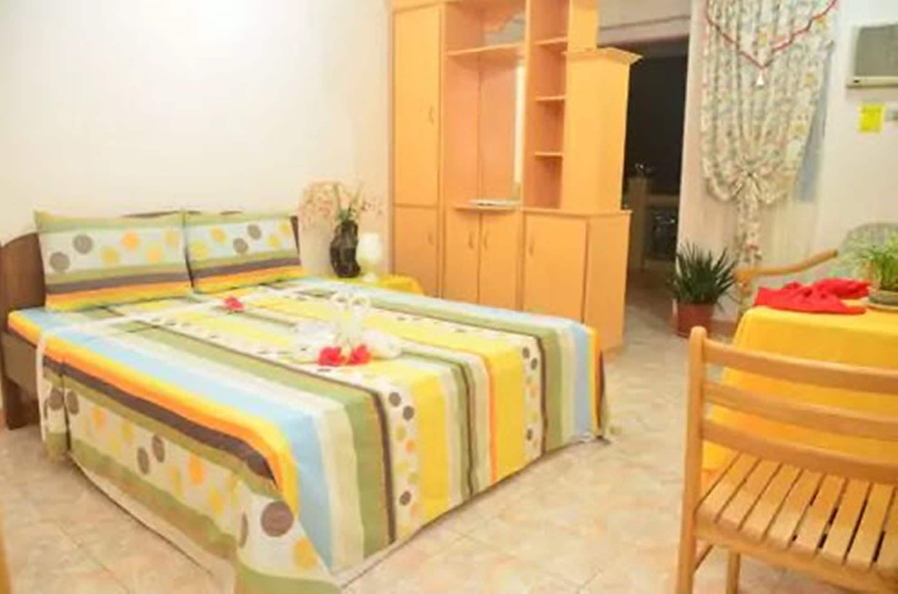 A bedroom with a bed, a wooden wardrobe, a small table with a chair, and a potted plant.