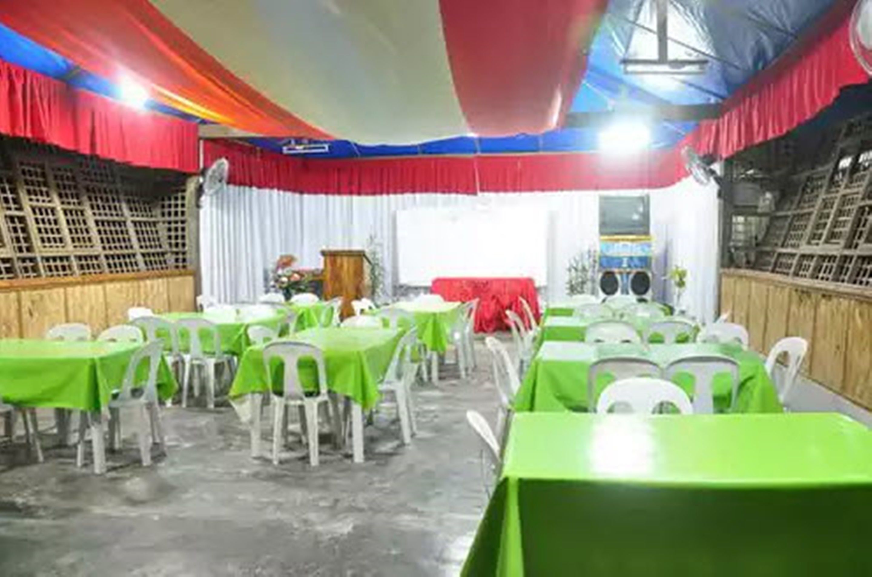 Una sala con varias mesas y sillas, algunas cubiertas con manteles verdes, y una zona de escenario con una cortina roja.