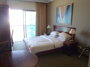 A hotel room with a bed, two bedside tables with lamps, a chair, a round table, a painting on the wall, and a view of the outside through a large window.