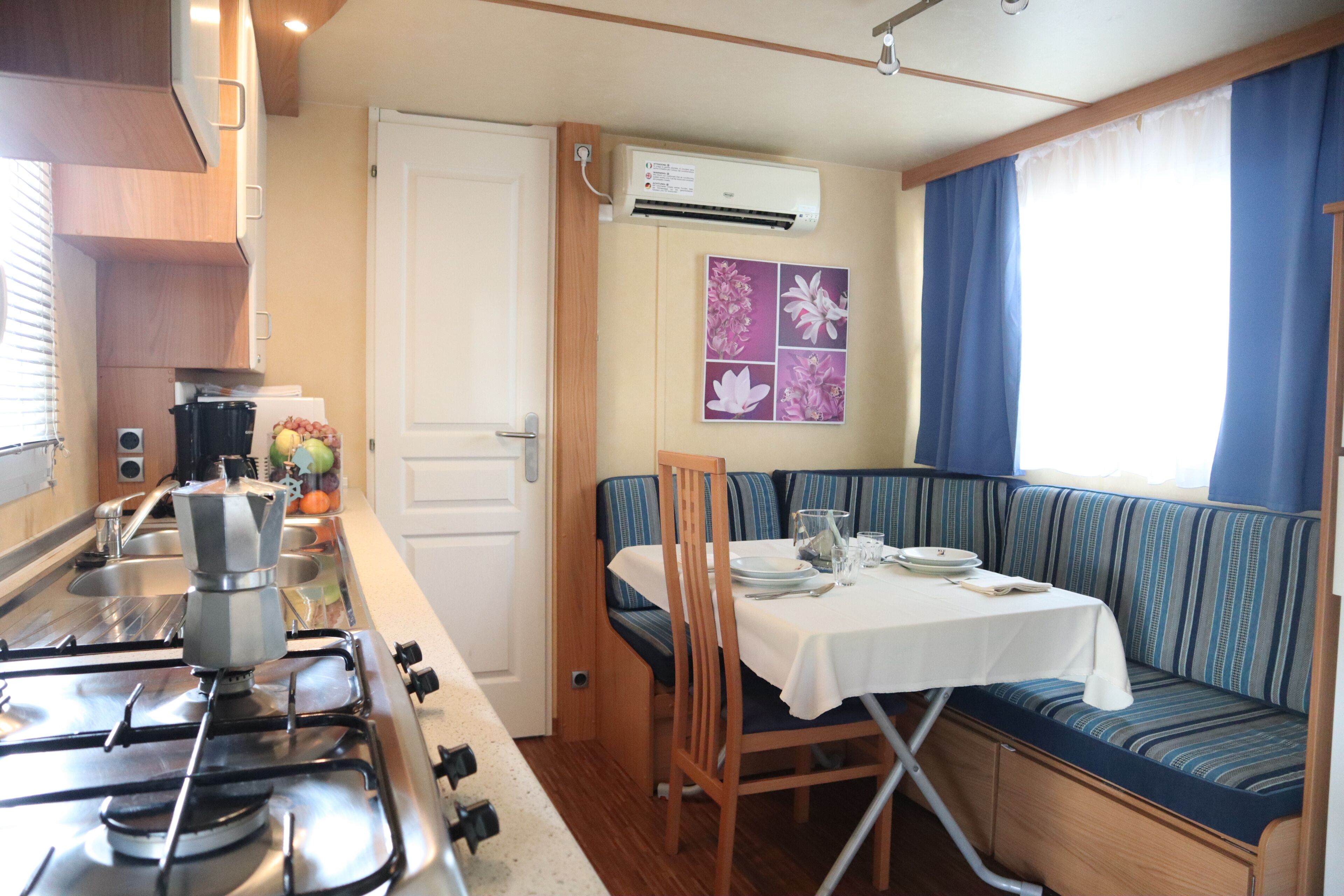 A compact kitchenette with a gas stove, a sink, and a small countertop. There is a dining area with a table and chairs, and a window with blue curtains.