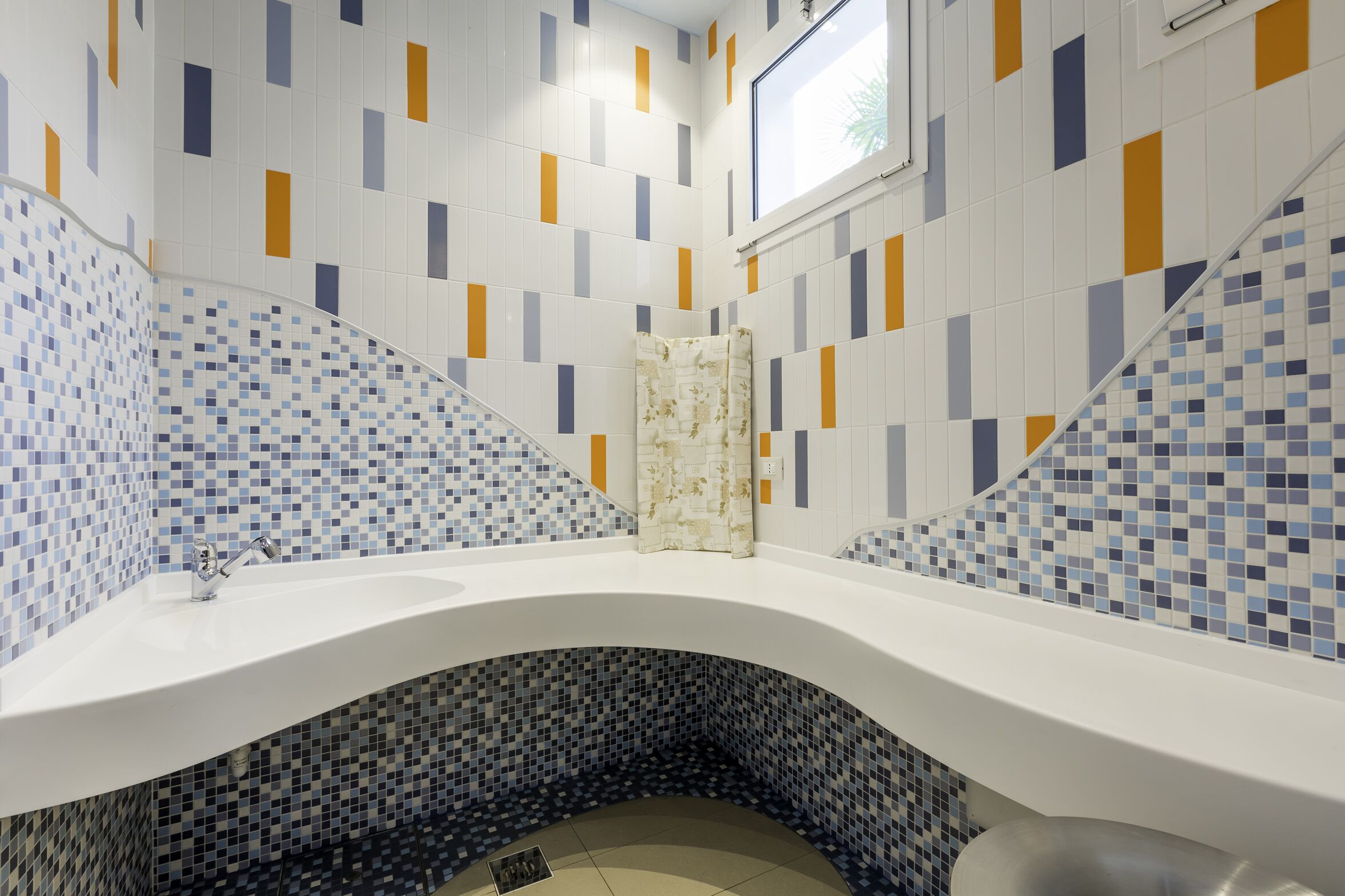 Un bagno con un lavandino bianco, pareti rivestite di piastrelle con motivi geometrici e una finestra sopra il lavandino.