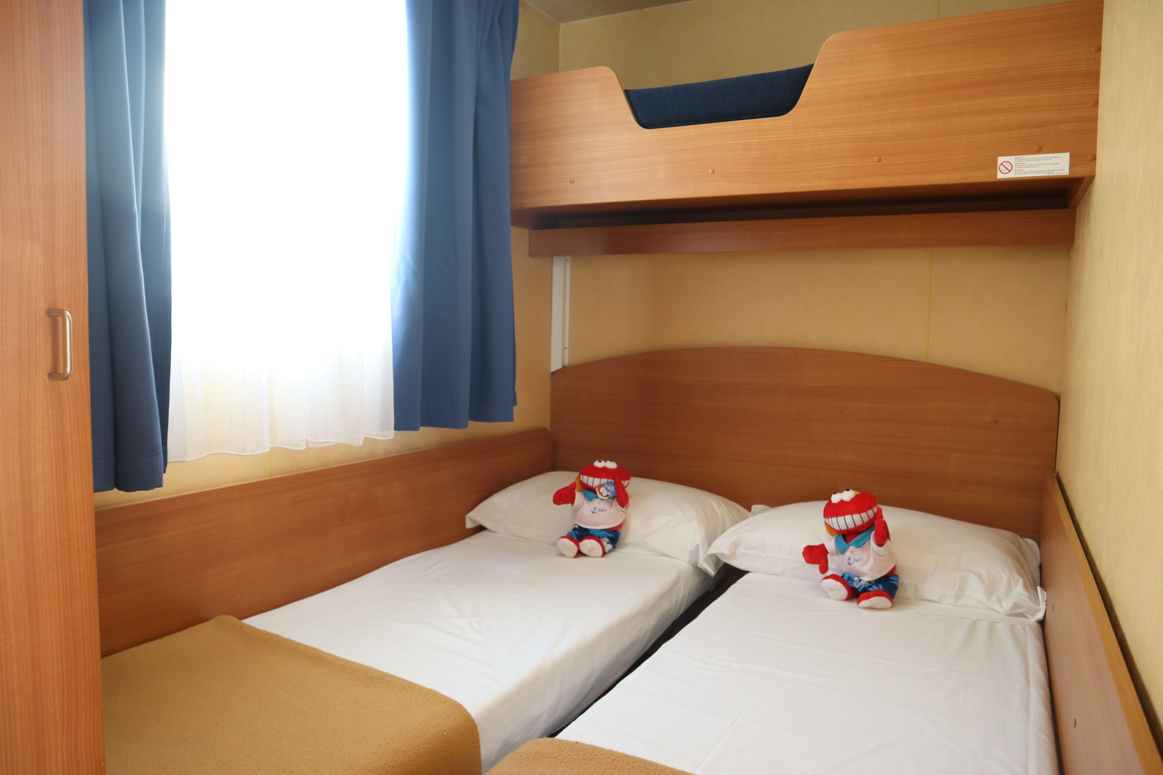Een kleine kamer met twee eenpersoonsbedden, een stapelbed erboven en een raam met gordijnen.