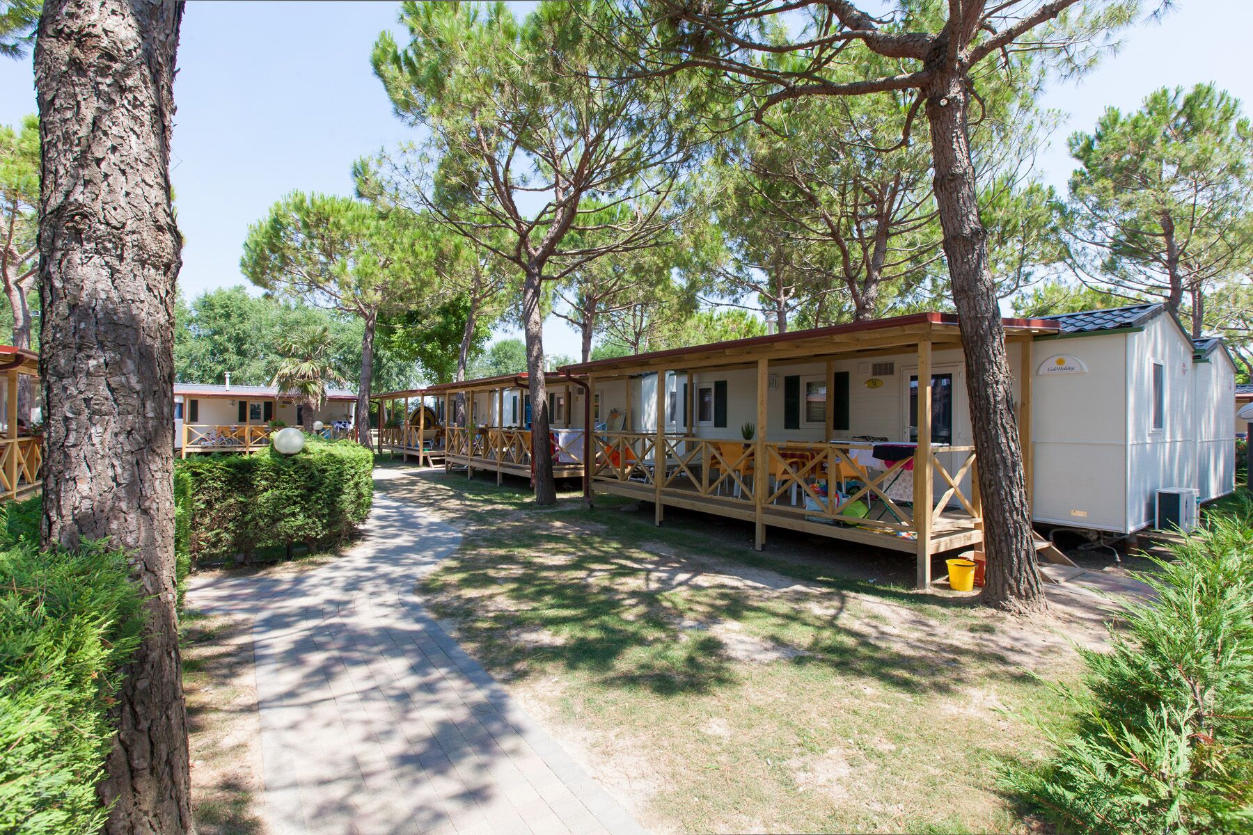 A row of mobile homes with wooden decks and outdoor seating areas, surrounded by trees and a paved path.