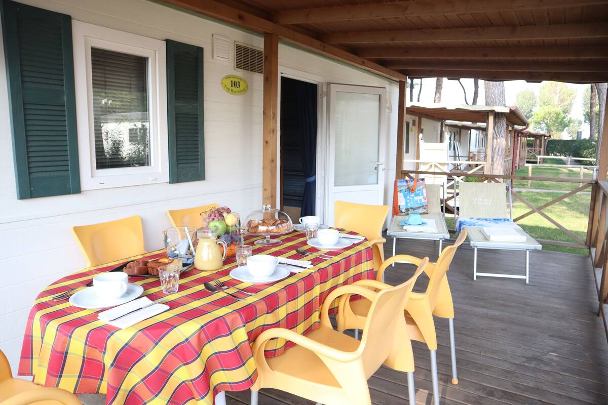 A covered outdoor dining area with a table set for four, yellow chairs, and a wooden deck.