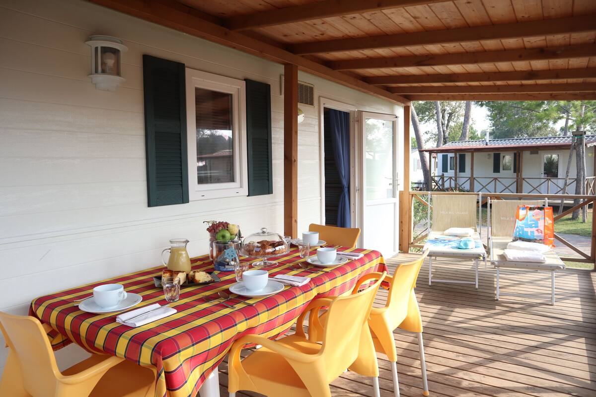 A covered outdoor dining area with a table set for two, yellow chairs, and a wooden deck.