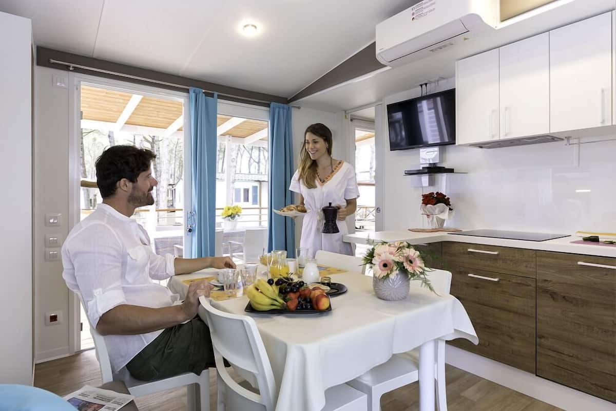 A modern kitchen with white cabinets, a dining table with a fruit platter, and a TV mounted above the counter.