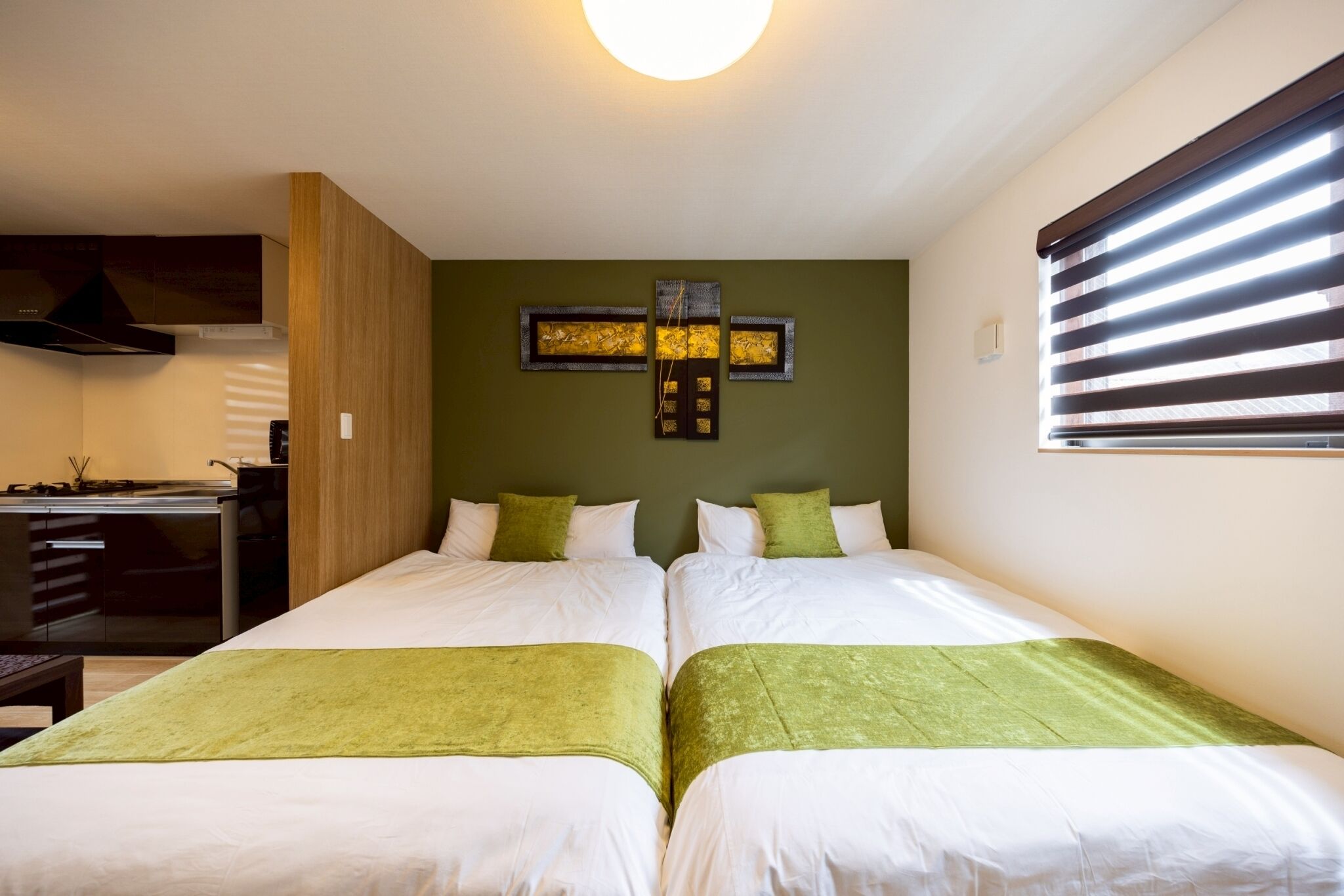 A hotel room with two beds, a kitchenette, and a large window with blinds.