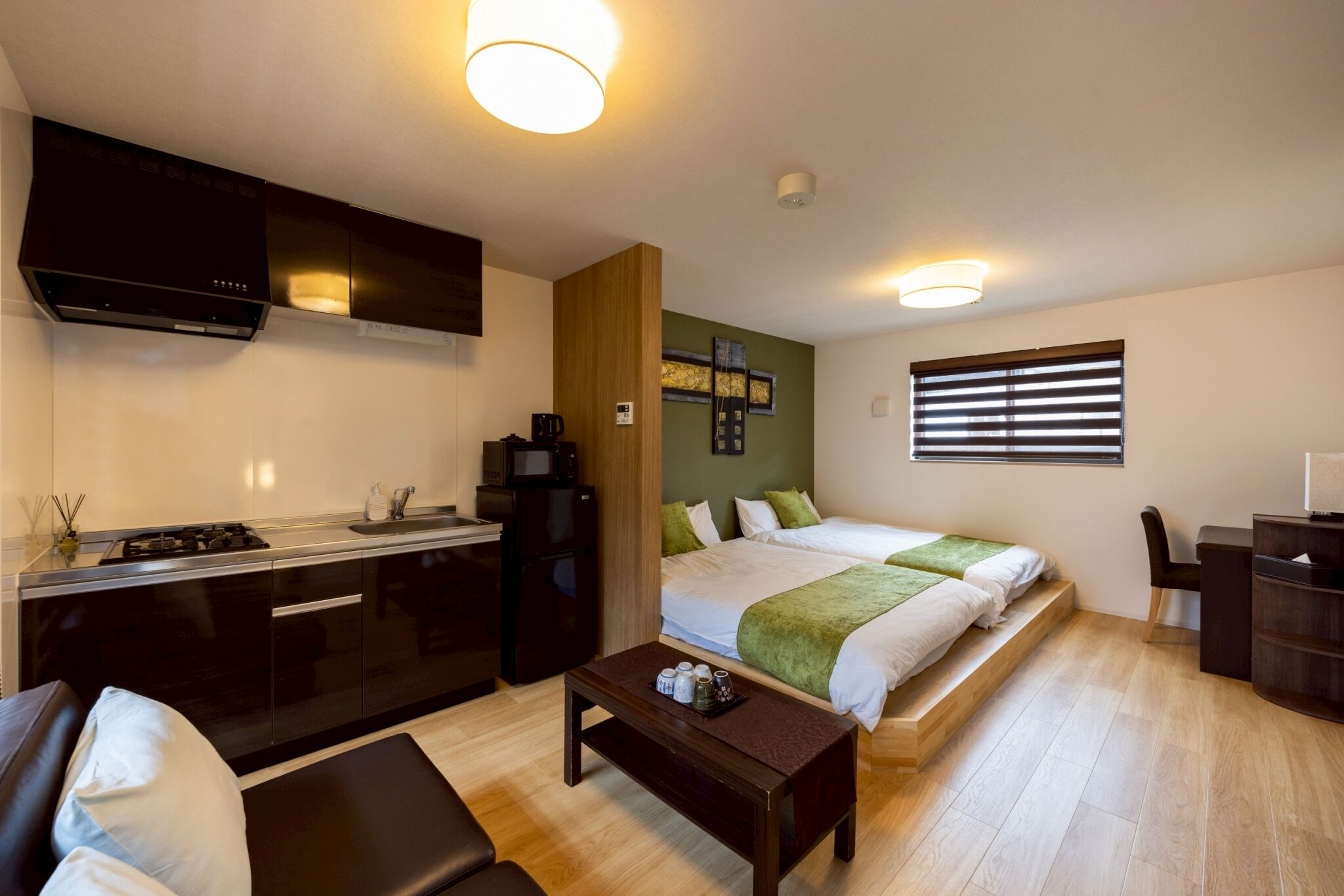 A compact hotel room with a kitchenette, a bed with green bedding, a desk, and a chair.