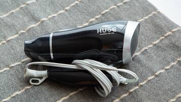 Ein schwarzer Haartrockner mit weißem Kabel und einer silbernen Düse, beschriftet mit „HUGE“, auf einer grauen Stoffoberfläche mit weißer Naht.