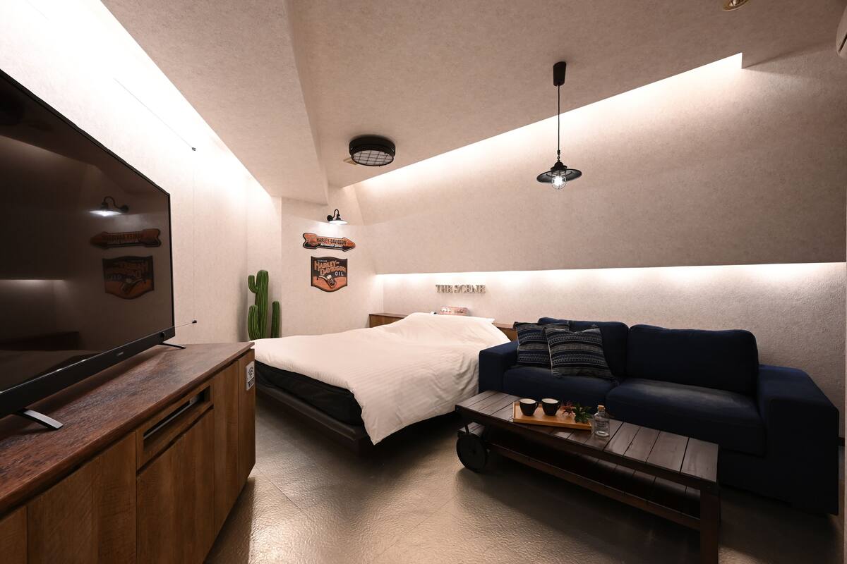 A compact room with a bed, a dark blue sofa, a wooden coffee table, a flat-screen TV, and a cactus.