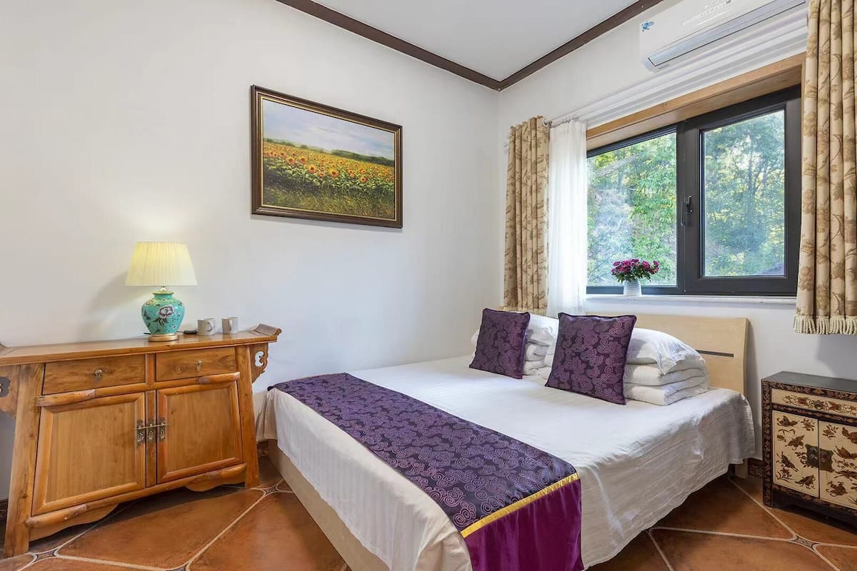 Un dormitori amb un llit, una petarrera de fusta, una làmpada, una finestra amb cortines i un quadre a la paret.