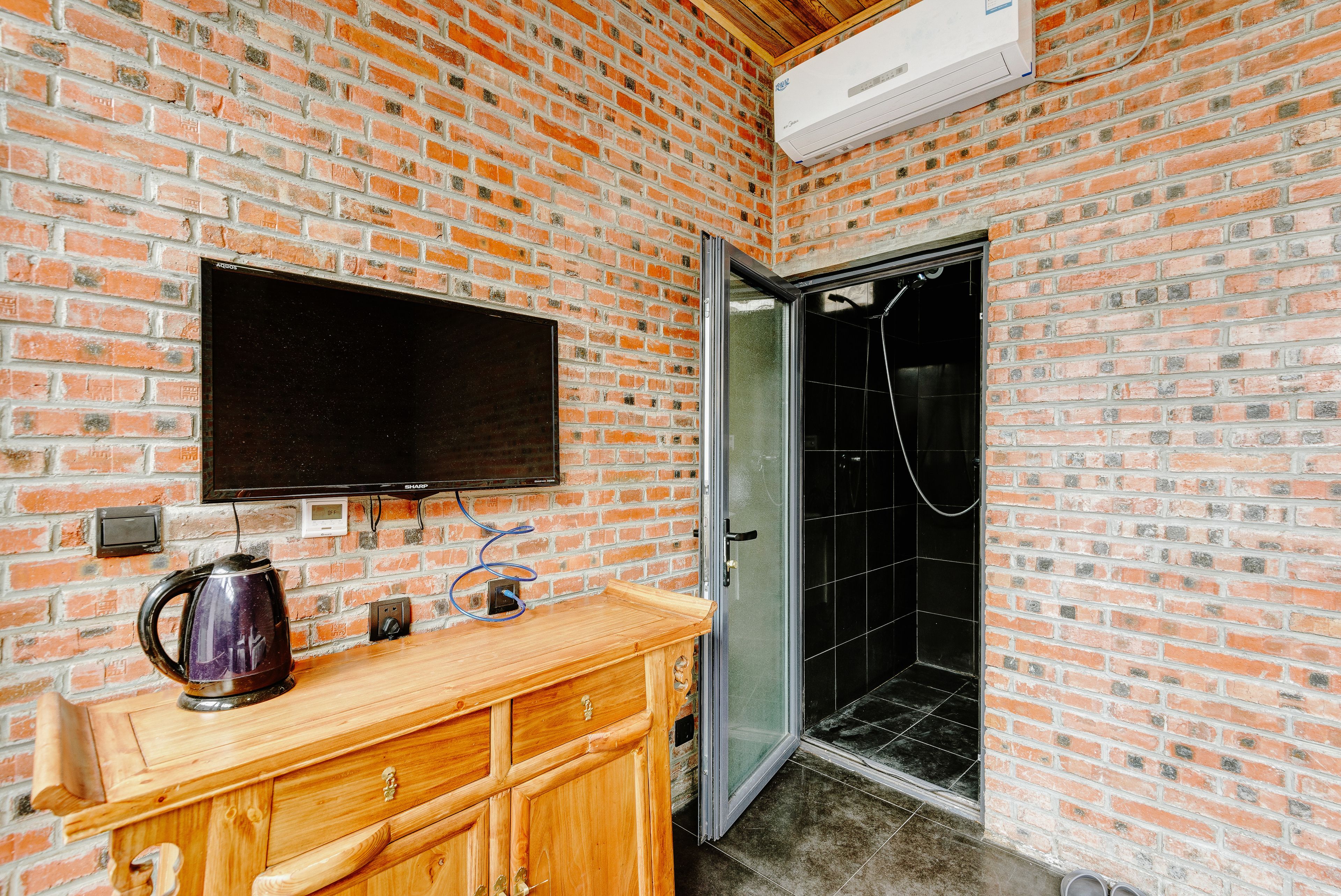 Un baño con pared de ladrillo, tocador de madera, tetera, televisor montado sobre el tocador y cabina de ducha de vidrio.