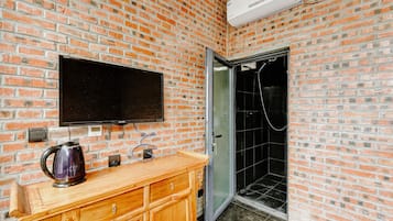 A bathroom with a brick wall, a wooden vanity, a kettle, a television mounted above the vanity, and a glass shower enclosure.